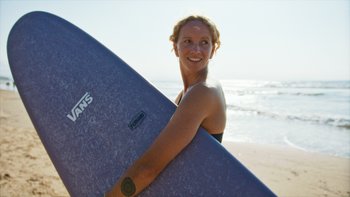 Eine Frau steht am Strand, ein Surfbrett unter dem Arm.