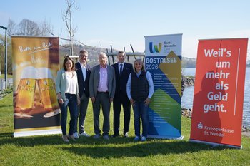 Vorstellung Veranstaltungsserie Bostalsee Fünf Personen stehen nebeneinander auf einer Wiese vor dem Bostalsee und posieren für ein Gruppenfoto. Im Hintergrund ist das Wasser des Sees zu sehen. Links und rechts sind drei große Roll-up-Banner aufgestellt: eines des Sponsors Bitburger, eines mit Informationen zur Veranstaltungsreihe und eines des Sponsors Kreissparkasse St. Wendel.