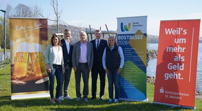 Fünf Personen stehen nebeneinander auf einer Wiese vor dem Bostalsee und posieren für ein Gruppenfoto. Im Hintergrund ist das Wasser des Sees zu sehen. Links und rechts sind drei große Roll-up-Banner aufgestellt: eines des Sponsors Bitburger, eines mit Informationen zur Veranstaltungsreihe und eines des Sponsors Kreissparkasse St. Wendel.